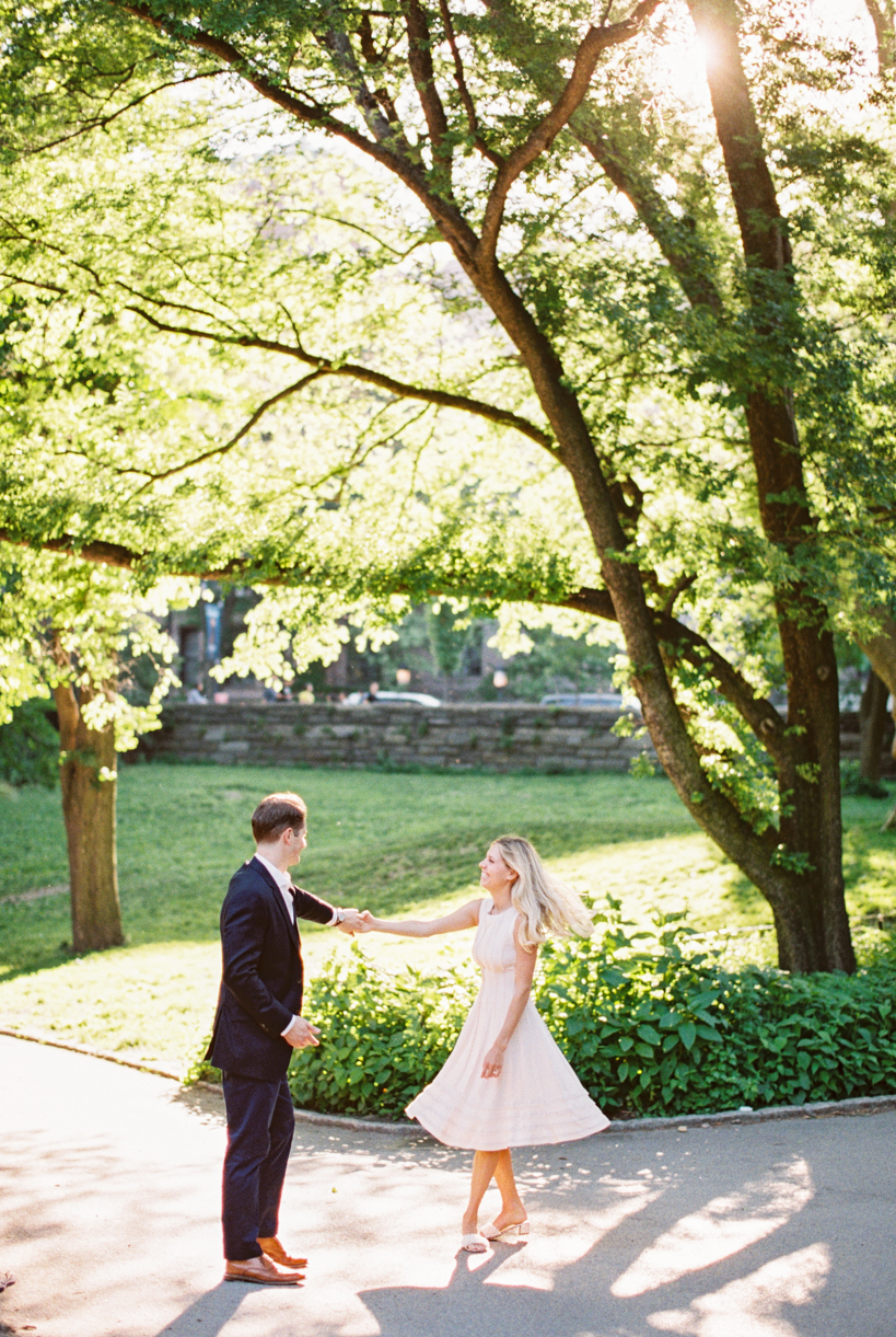 guy twirls his fiance wearing short ivory dress in Central Park during NYC Engagement Session in Central Park