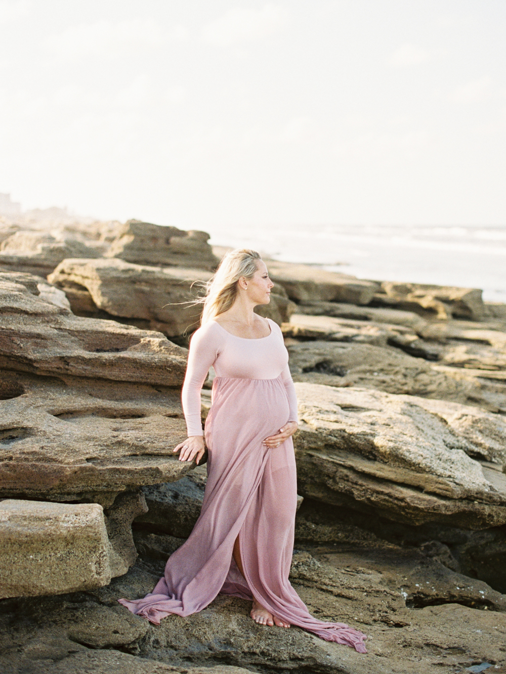mother to be poses on rocky beach in long pink dress for Maternity and Newborn Sessions